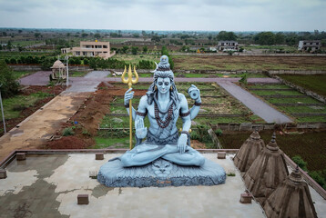 Lord Shiva statue aerial view, Mahadev sculpture with trident, Shiva with snake and trident landscape, Peaceful Shiva idol countryside view, Tranquil Lord Shiva outdoor statue.





