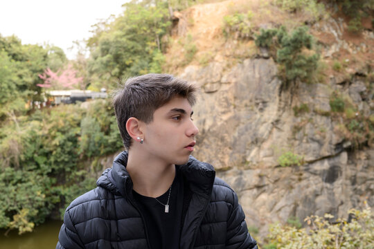 Jovem pensativo ao ar livre, com jaqueta preta, em frente a pared&atilde;o de pedras e vegeta&ccedil;&atilde;o. Retrato urbano-natural ideal para temas de juventude, atitude e natureza.
