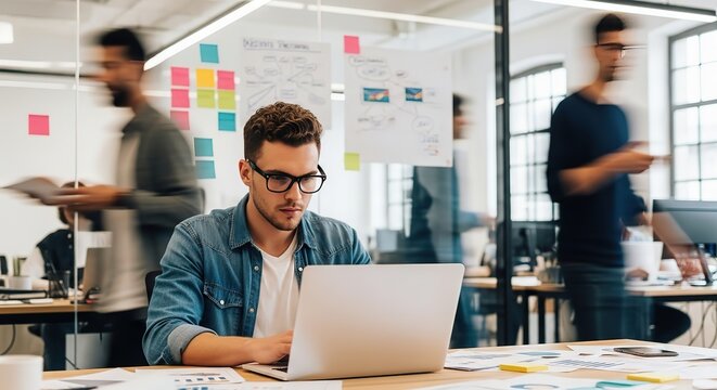 Focused young professional working diligently on a laptop in a modern bustling office environment with colleagues blurred in motion