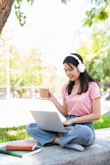 Young female student with laptop and headphones studying online sitting outdoor drinking coffee at...