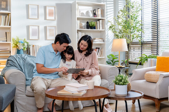 Asian Family time.Happy asian mom,dad and daughter using digital tablet,learning,searching,browsing internet online at home,sitting together on couch indoors.Gadgets and modern lifestyle concept