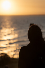 Fototapeta premium Woman taking photo of golden sunset over ocean with phone