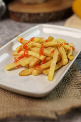 Close-up of crispy golden french fries topped with a drizzle of ketchup, served on a white rectangular plate over a burlap placemat.