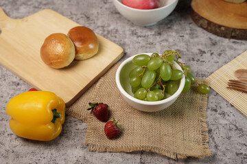 A healthy food flat lay with fresh green grapes in a bowl, strawberries, a yellow bell pepper, and bread rolls on a rustic grey textured background.