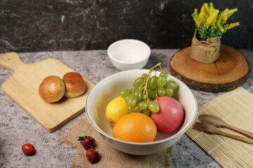 A vibrant bowl of fresh fruits including grapes, a lemon, an orange, and an apple, with bread and strawberries on a grey background.