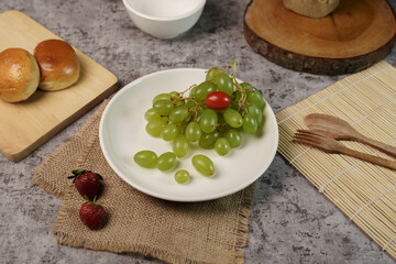Food composition flat lay featuring green grapes and a cherry tomato on a plate, with bread and strawberries on a grey background.