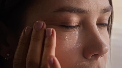 Close-up of young beautiful woman applying hydrating gel on perfect skin of her face and looking at herself in mirror while tapping cosmetic product with fingertips - Powered by Adobe