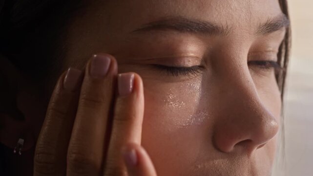 Close-up of young beautiful woman applying hydrating gel on perfect skin of her face and looking at herself in mirror while tapping cosmetic product with fingertips