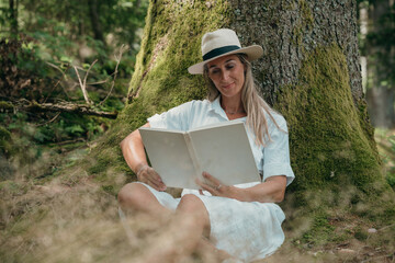 A peaceful forest day captured: blonde woman in summer dress leans on tree wearing straw hat...