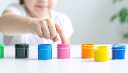 Young Child Dipping Finger Into Pink Paint From An Array Of Brightly Colored Paint Jars On A White Tabletop
