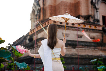 Woman in traditional Thai dress holding a white umbrella and touching a lotus flower at Wat Chedi...