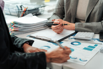 Two businesswomen engaged in a discussion about financial charts and documents at an office desk, analyzing business growth trends and diligently taking notes for future strategies