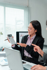 Two Asian businesswomen are analyzing financial data using a calculator and laptop, discussing...