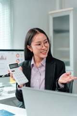 Asian businesswoman wearing glasses and a tailored suit, holding a calculator while discussing...