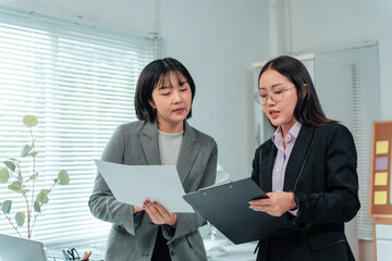 Two Asian businesswomen in tailored suits are standing in a modern office, comparing documents and...