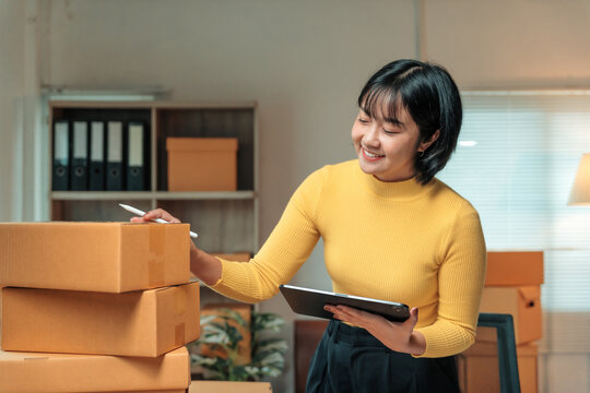 Smiling young Asian woman entrepreneur checking cardboard boxes and using digital tablet while working in home office, managing online orders and preparing for shipping