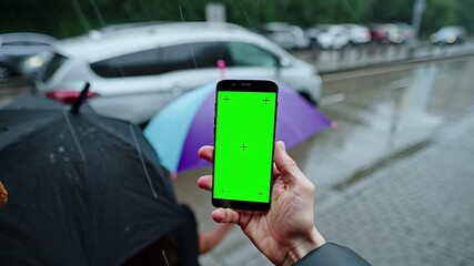 A hand holds a smartphone with a green screen under an umbrella on a rainy city street