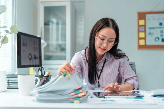 Young professional woman reviewing documents and using tablet, efficiently handling administrative tasks and financial analysis in a modern office workspace - Powered by Adobe