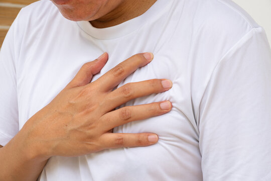 In a softly lit, neutral setting, an Asian man in a white shirt touches his chest with concern, suggesting possible discomfort or heart-related symptoms. Healthcare and medical concept.