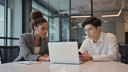 Black adult woman Asian adult man business professionals collaborate discuss project laptop smartphone modern office workspace meeting teamwork strategy data review