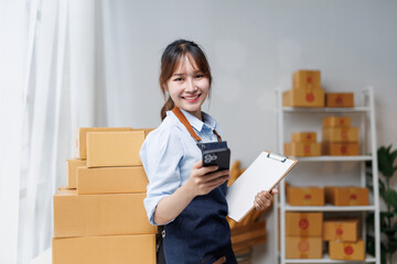 Cheerful young Asian female seller managing inventory with a mobile phone and clipboard in a...