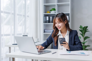 Young Asian businesswoman working diligently on a laptop while managing her smartphone, balancing tasks in a modern office environment, embodying the spirit of entrepreneurship and success
