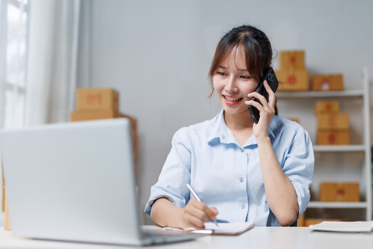 Young Asian woman entrepreneur, small business owner talking on mobile phone while working with laptop computer receiving online orders and taking notes in a notebook at home office