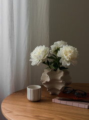 A bouquet of white peonies, a cup of coffee, a notebook on a round wooden table in the living room