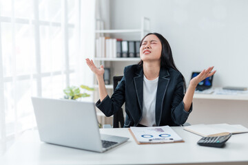 Young Asian businesswoman raising hands with frustrated expression while working on laptop at...