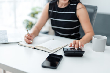 Businesswoman is calculating and taking notes, performing accounting and financial analysis using calculator, laptop and smartphone at her workplace