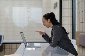 Asian businesswoman is pointing at her laptop and laughing while making a video call in a modern office environment, enjoying a moment of amusement during her workday