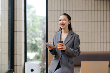Smiling businesswoman using a mobile phone while sipping takeaway coffee and gazing out of a modern...