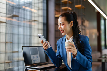 Smiling businesswoman in a blue suit, enjoying tea while using a mobile phone during a work break, with a laptop displaying charts and data analysis in the background