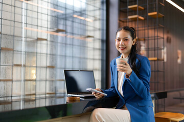 Young Asian businesswoman sipping coffee and using a smartphone while working on a laptop in a...