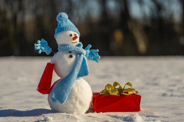 Snowman with Christmas gift in a snowy field. Funny snowman with a carrot nose and scarf and winter hat. Snowman decorated for Christmas. Snowman for Christmas card design.