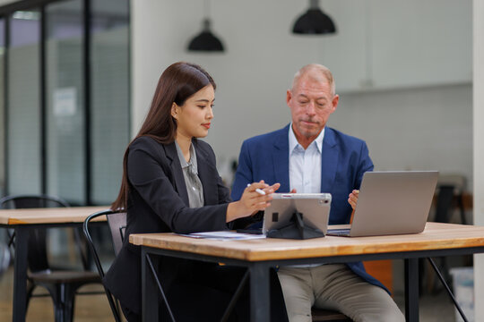 Mature manager mentor talking to Asian female coworker, showing online project results at Business partners in meeting. Two happy diverse professional executives team working on laptop in office 