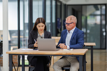 Mature manager mentor talking to Asian female coworker, showing online project results at Business partners in meeting. Two happy diverse professional executives team working on laptop in office