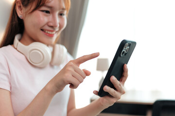 Happy young woman with headphones around her neck is using smartphone at home, tapping on screen and smiling while browsing internet or social media