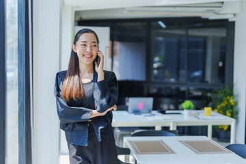 Asian businesswoman standing by a window in a modern office, smiling while talking on her mobile phone and looking away, exuding confidence and professionalism in her elegant suit