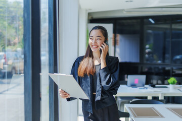 Asian businesswoman standing by a window, engaging in a phone conversation while holding a clipboard, smiling and glancing away, working diligently in a modern office environment