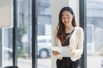 Young Asian businesswoman smiling and taking notes on a clipboard while standing next to a window in her office, she's wearing a white jacket and black trousers