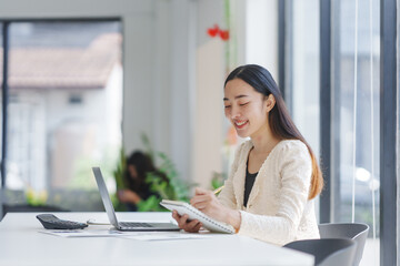 Young Asian businesswoman smiling and taking notes in her notebook while working on a laptop in a modern office, enjoying her productive workday