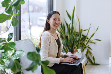 Young Asian businesswoman sitting comfortably on a modern chair, working intently on her laptop in a stylish office filled with plants, smiling confidently and radiating satisfaction