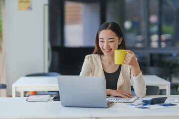 Young happy professional woman is drinking from a yellow mug and using laptop at workplace, smiling and enjoying her break time in a modern office