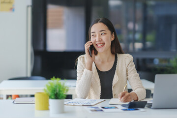 Smiling businesswoman talking on phone and taking notes while sitting at desk in modern office, working with financial documents and using laptop