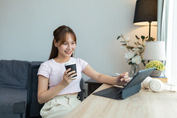 Happy young Asian businesswoman enjoying coffee while working from home, using a tablet and stylus at her cozy desk in the living room, surrounded by natural light and plants