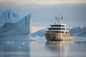 Expedition yacht navigates icy waters alongside a colossal iceberg under a soft hazy sky.