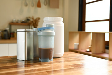 A jar of protein powder sits on a wooden table next to a blender and a canister. The scene suggests that someone is preparing a healthy meal or snack. The presence of the blender