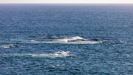 Fototapeta premium A dynamic ocean scene with waves crashing over submerged rocks, sending sea spray into the air under clear skies. Perfect for travel, nature, and seascape themes.