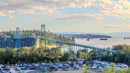 Panoramic view of the iconic Lions Gate Bridge spanning the inlet, with cargo ships, forested hills, and a residential neighborhood below at sunset.